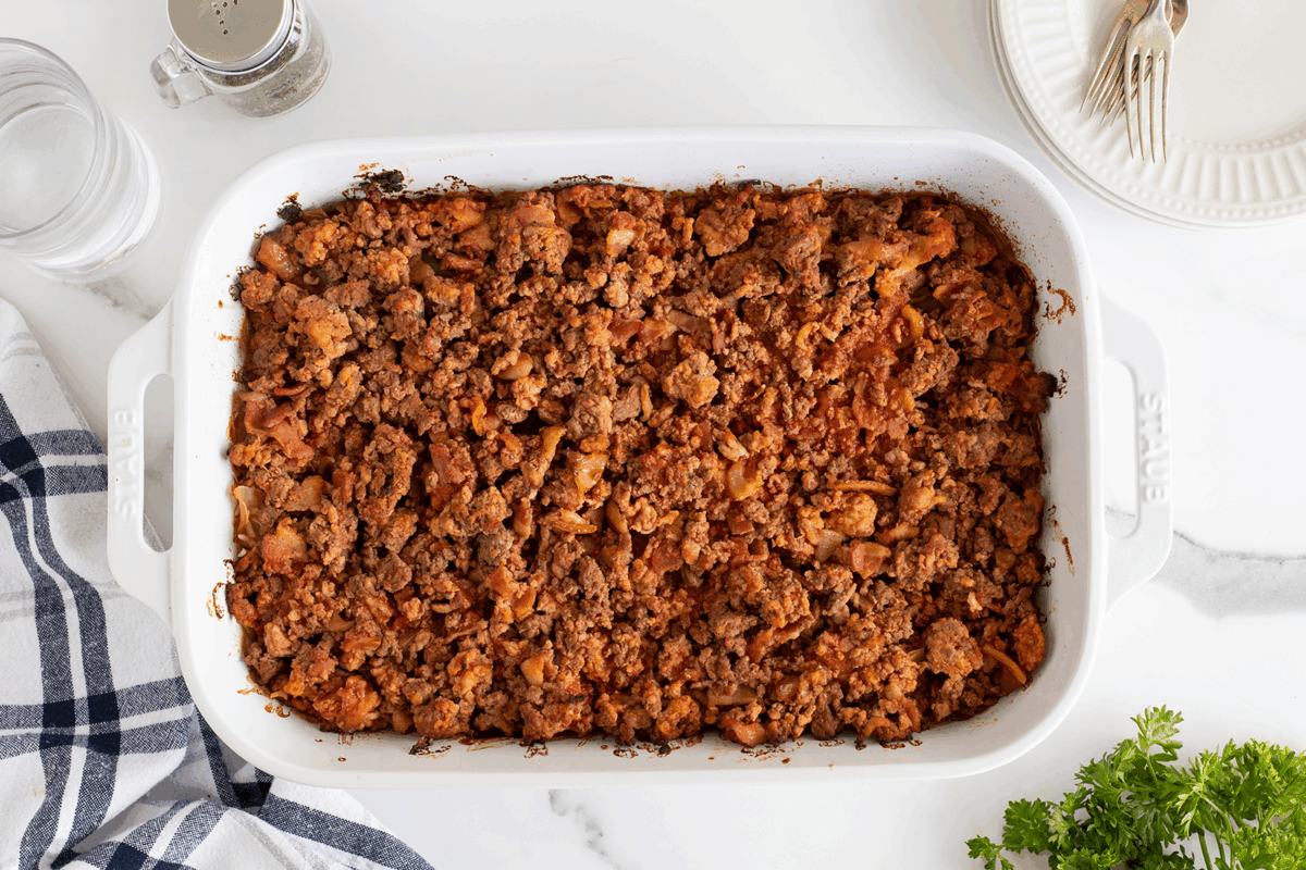 A white baking dish filled with baked ground beef and tomato casserole sits on a white countertop, surrounded by a striped towel, a glass of water, a pepper shaker, stacked plates with forks, and a bunch of fresh parsley.