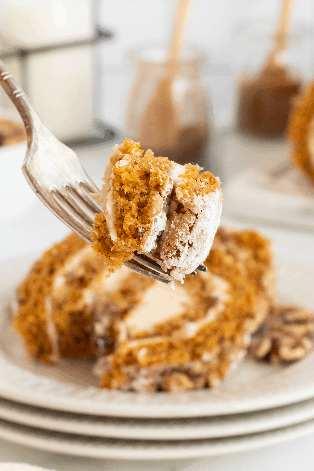 A close-up of a fork holding a bite of pumpkin roll cake with cream cheese filling, with a slice of the cake on a plate in the background. The cake is rolled and dusted with powdered sugar.