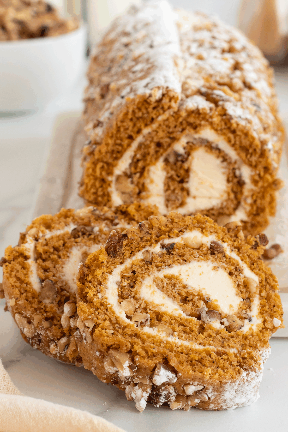 A close-up of a sliced pumpkin roll cake with a creamy white filling, topped with powdered sugar and chopped pecans, displayed on a white surface.