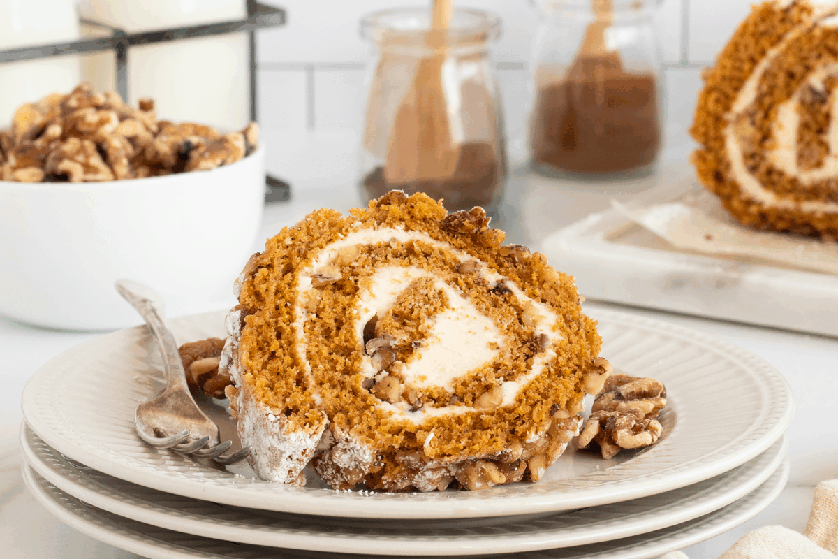 A slice of pumpkin roll cake with cream cheese filling sits on a white plate, garnished with chopped walnuts; a fork is beside the cake. In the background are jars of cinnamon and a bowl of walnuts.