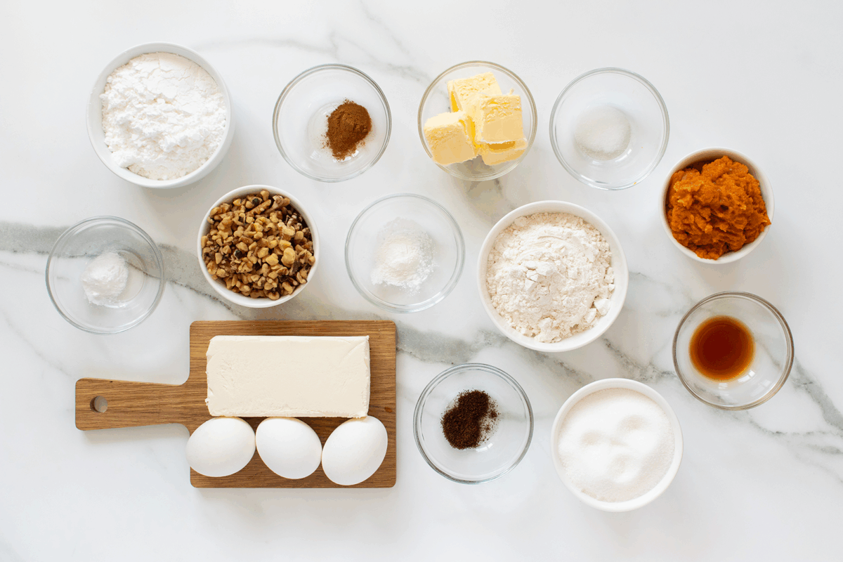 A variety of baking ingredients are arranged on a white marble surface, including eggs, butter, flour, sugar, nuts, spices, baking powder, baking soda, pumpkin puree, and vanilla extract in small bowls and dishes.