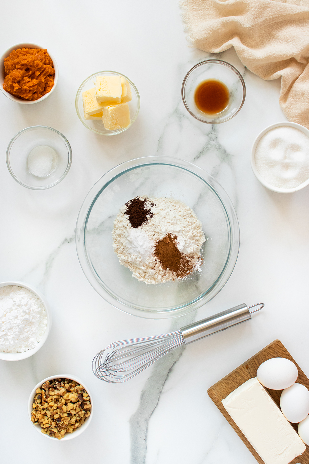 A glass bowl with flour and spices sits on a marble countertop surrounded by eggs, butter, cream cheese, sugar, vanilla, chopped nuts, pumpkin puree, baking powder, and a metal whisk.