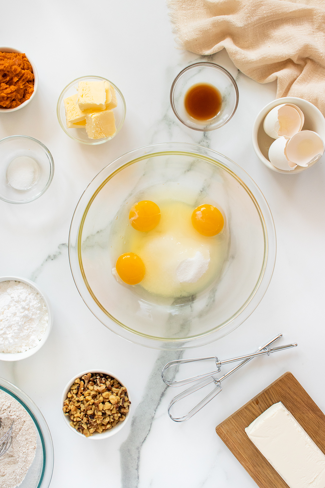 A glass bowl with eggs and sugar sits on a marble counter, surrounded by bowls of ingredients like butter, flour, sugar, nuts, and pumpkin puree, along with a hand mixer, vanilla, and cracked egg shells.