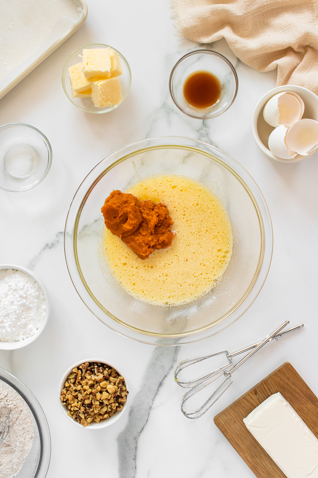 A mixing bowl with eggs and pumpkin puree, surrounded by baking ingredients: chopped nuts, powdered sugar, butter, vanilla, flour, salt, cream cheese, and cracked eggs on a marble countertop with a hand mixer nearby.