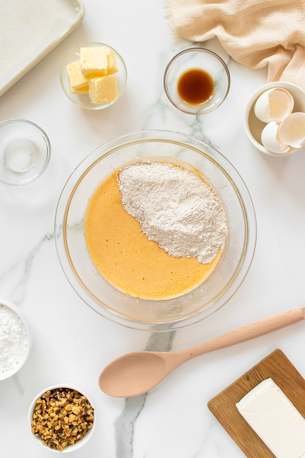 A glass bowl with batter and flour sits on a marble countertop, surrounded by butter, vanilla, chopped nuts, eggshells, a wooden spoon, cream cheese, and small bowls of sugar and salt.
