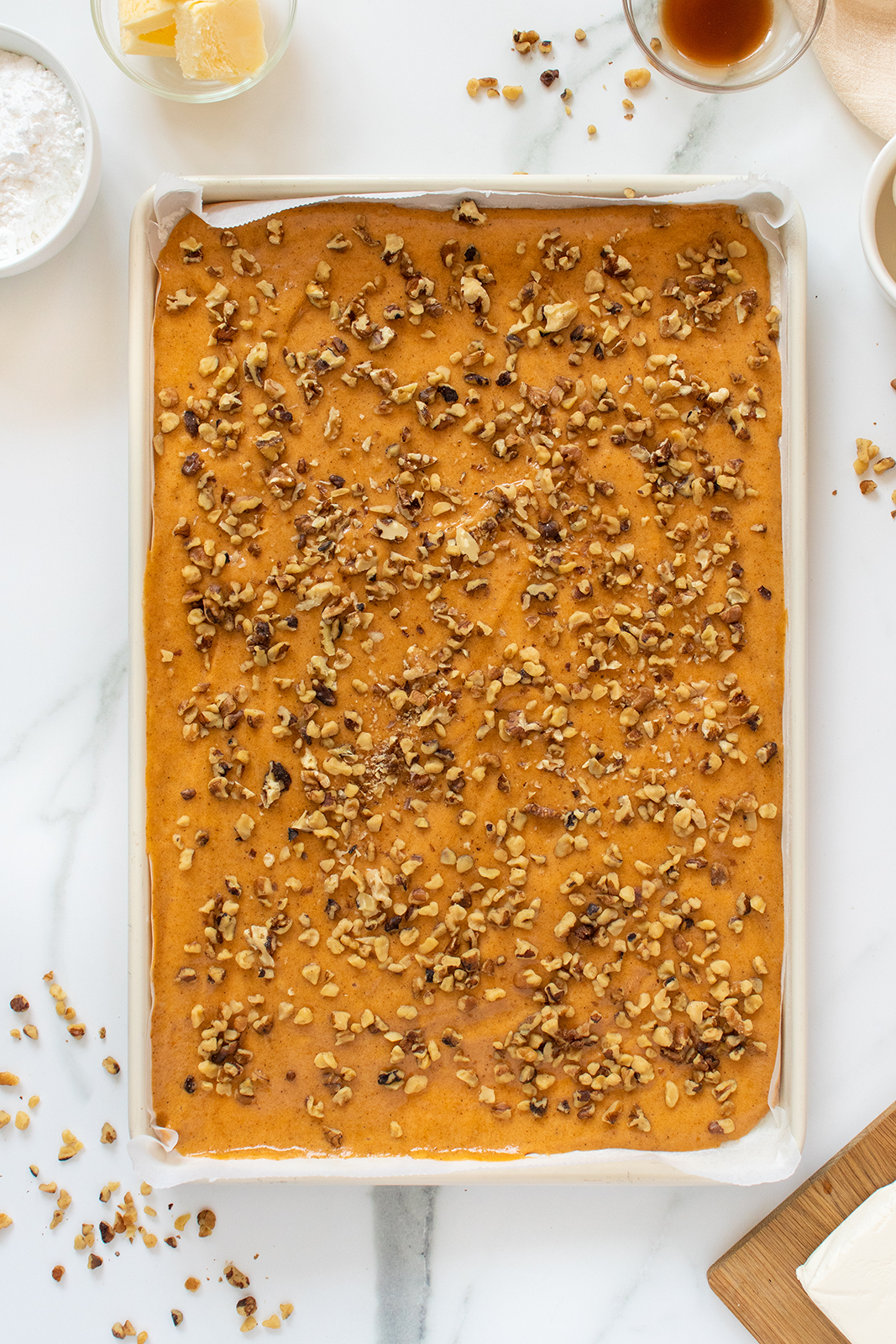 A rectangular sheet cake topped with chopped nuts sits on parchment paper, surrounded by baking ingredients like butter, flour, and brown sugar on a white marble surface.