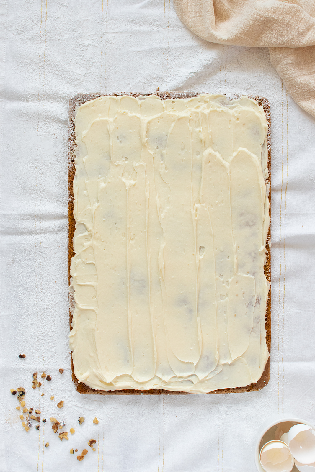 A rectangular sheet cake covered with smooth white icing sits on a white cloth. Eggshells, nuts, and a beige napkin are nearby. The scene is bright and minimalistic.