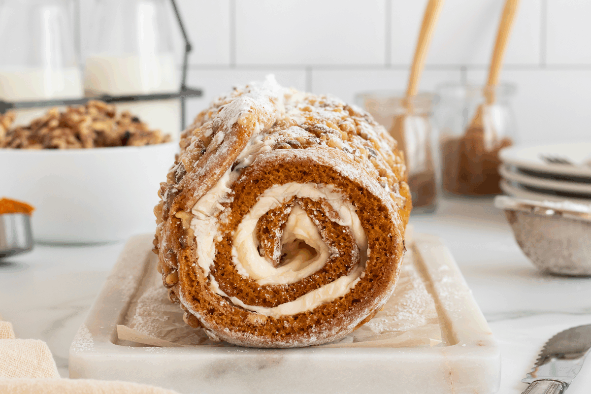 A close-up of a pumpkin roll cake with cream cheese filling, topped with powdered sugar and chopped nuts, displayed on a parchment-lined marble board in a bright kitchen setting.