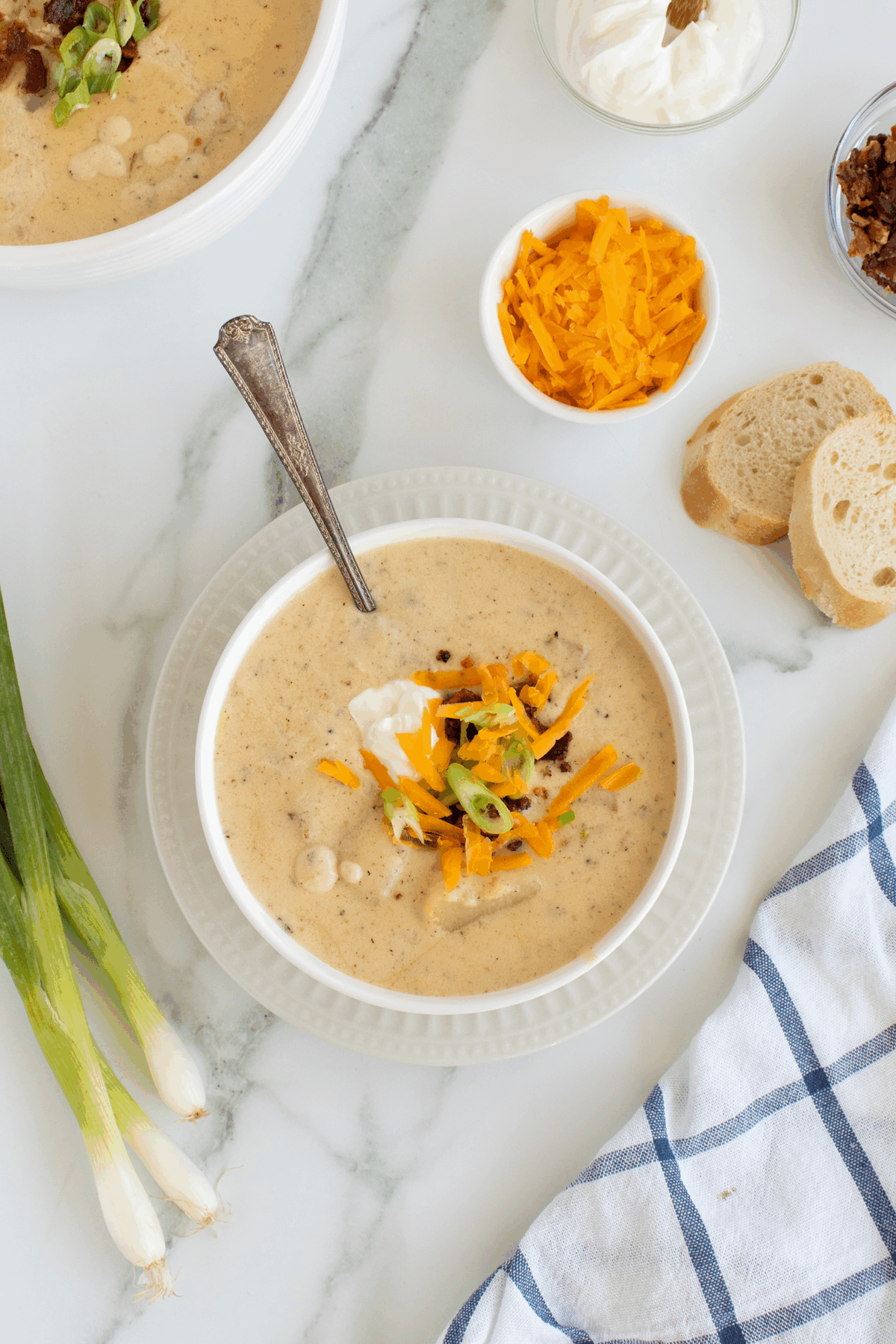 A bowl of creamy soup topped with shredded cheddar cheese, sour cream, and sliced green onions sits on a plate with a spoon. Nearby are sliced bread, grated cheese, bacon bits, green onions, and a blue plaid napkin.