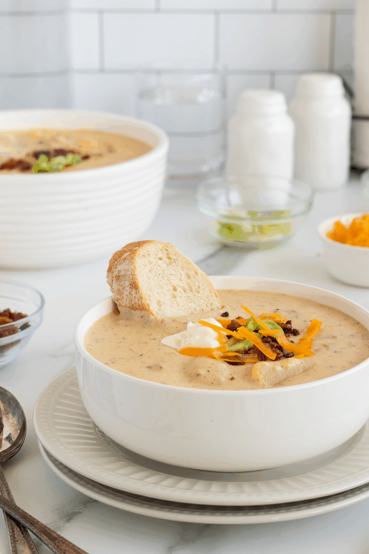 A bowl of creamy soup topped with shredded cheese, bacon bits, green onions, and a dollop of sour cream, served with a slice of bread. Additional toppings and another bowl of soup are in the background.