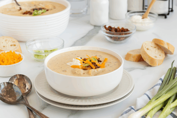 A bowl of creamy soup topped with shredded cheese, bacon bits, and green onions, served with sliced baguette on a plate. Nearby are bowls of cheese, green onions, raisins, and sour cream.
