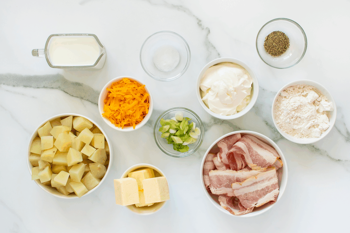 A top-down view of ingredients on a white surface, including diced potatoes, butter, bacon, chopped green onions, shredded cheddar cheese, flour, sour cream, milk, salt, and pepper.