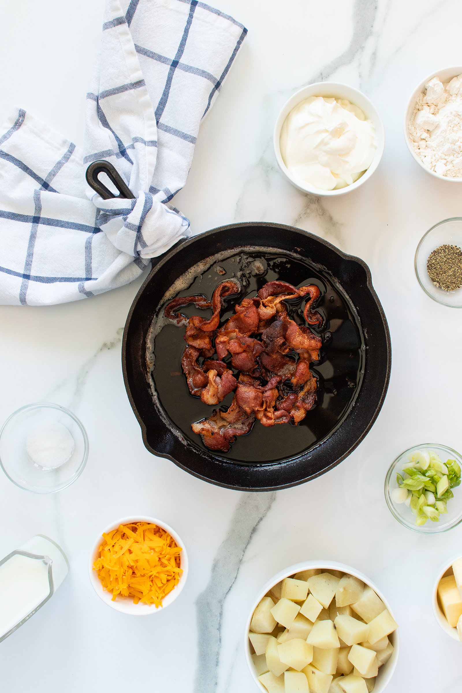 A skillet with cooked bacon sits in the center, surrounded by bowls of diced potatoes, shredded cheddar, chopped green onions, milk, sour cream, flour, pepper, and a striped kitchen towel on a marble counter.