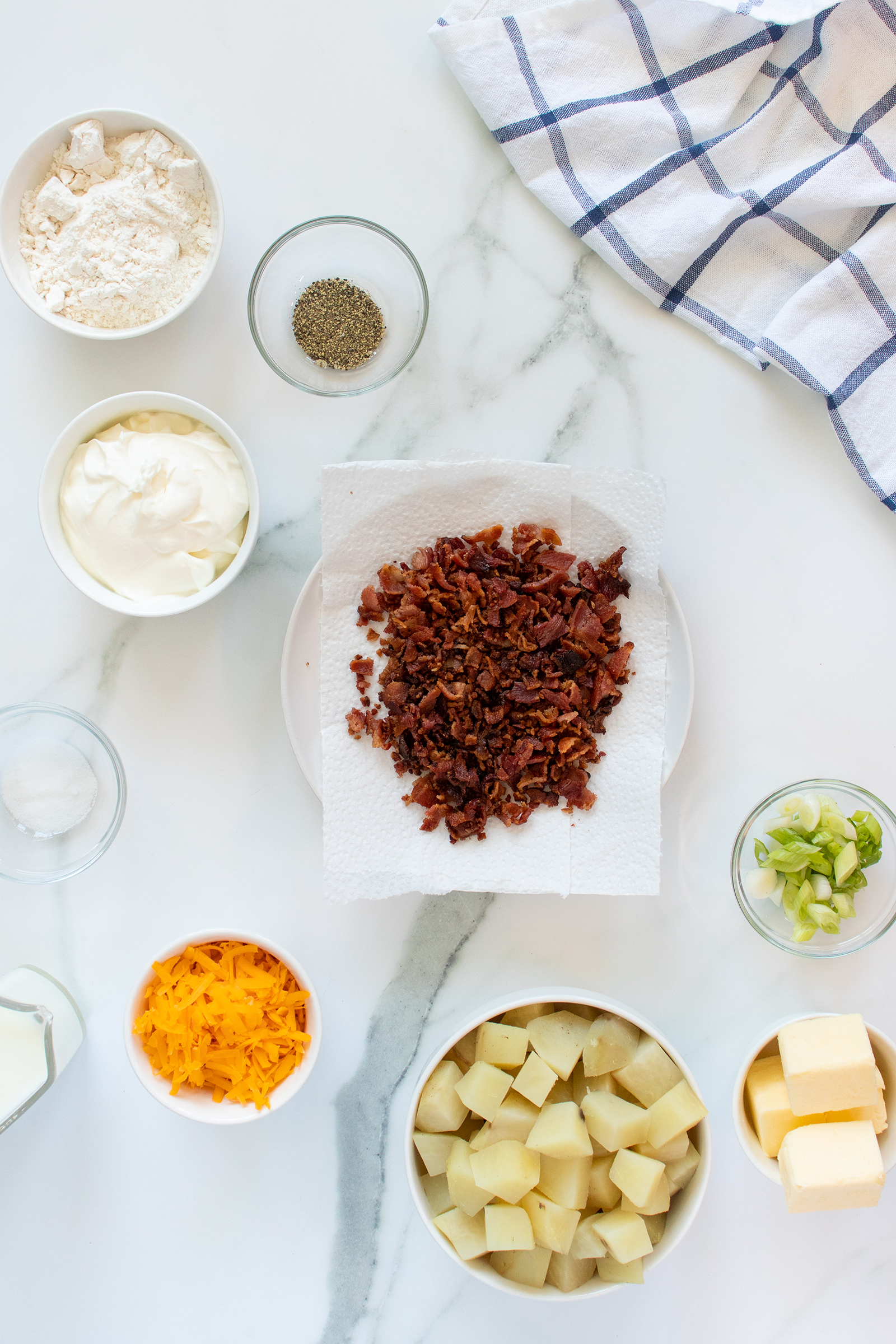 A top-down view of small bowls containing chopped cooked bacon, cubed potatoes, shredded cheddar cheese, sliced green onions, butter, flour, mayonnaise, salt, and pepper, arranged on a white marble surface with a checkered towel.