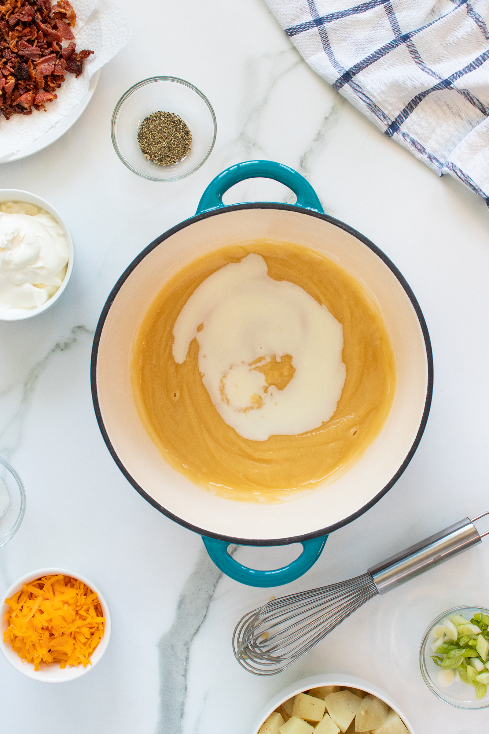 A blue-handled pot with a light brown roux and milk being added sits on a marble counter, surrounded by bowls of shredded cheese, bacon, sour cream, green onions, pepper, and a whisk.