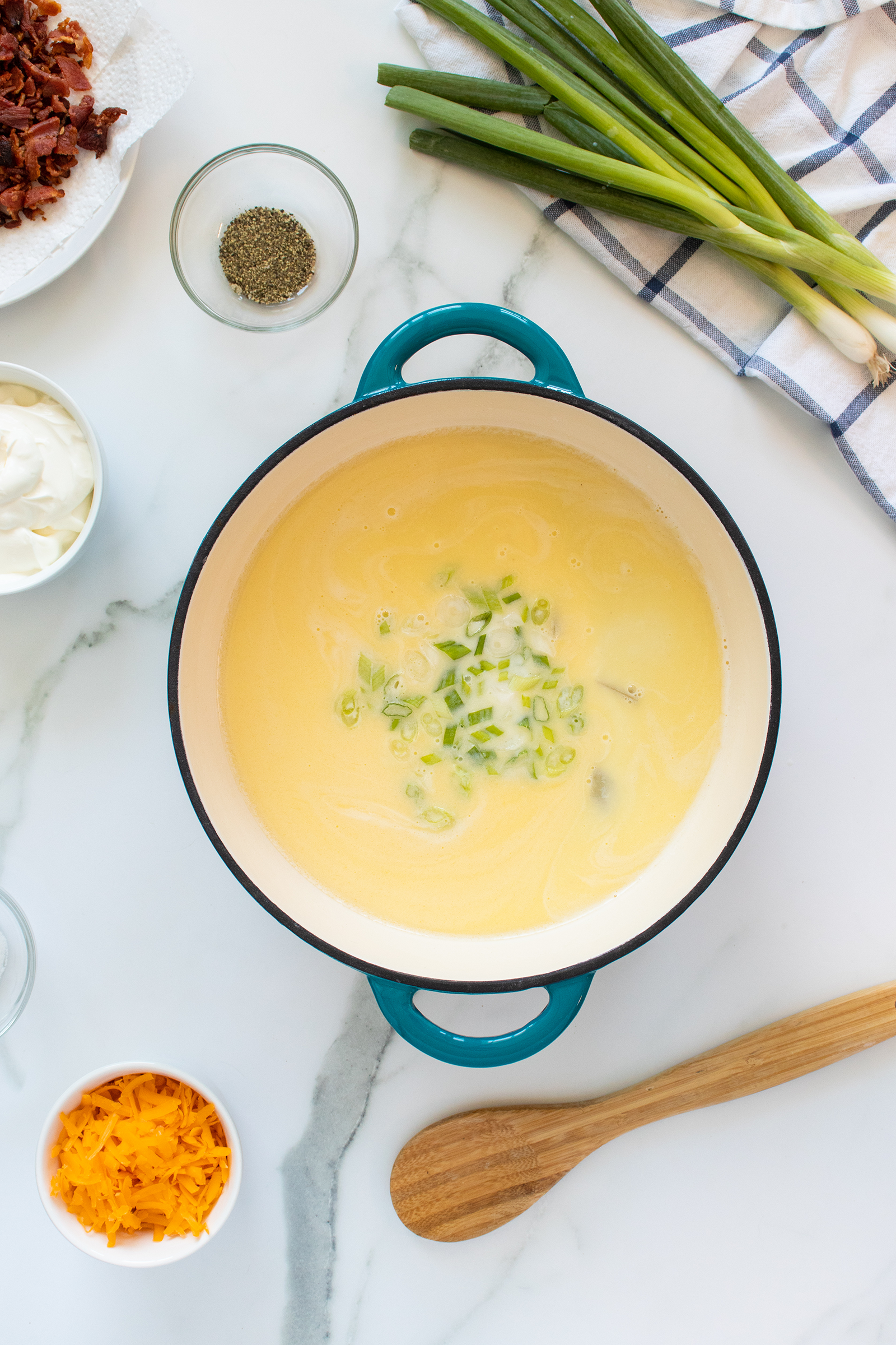 A pot of creamy soup topped with chopped green onions sits on a marble surface. Surrounding it are bowls of shredded cheese, sour cream, crumbled bacon, black pepper, and a bunch of green onions. A wooden spoon and a checkered cloth are nearby.