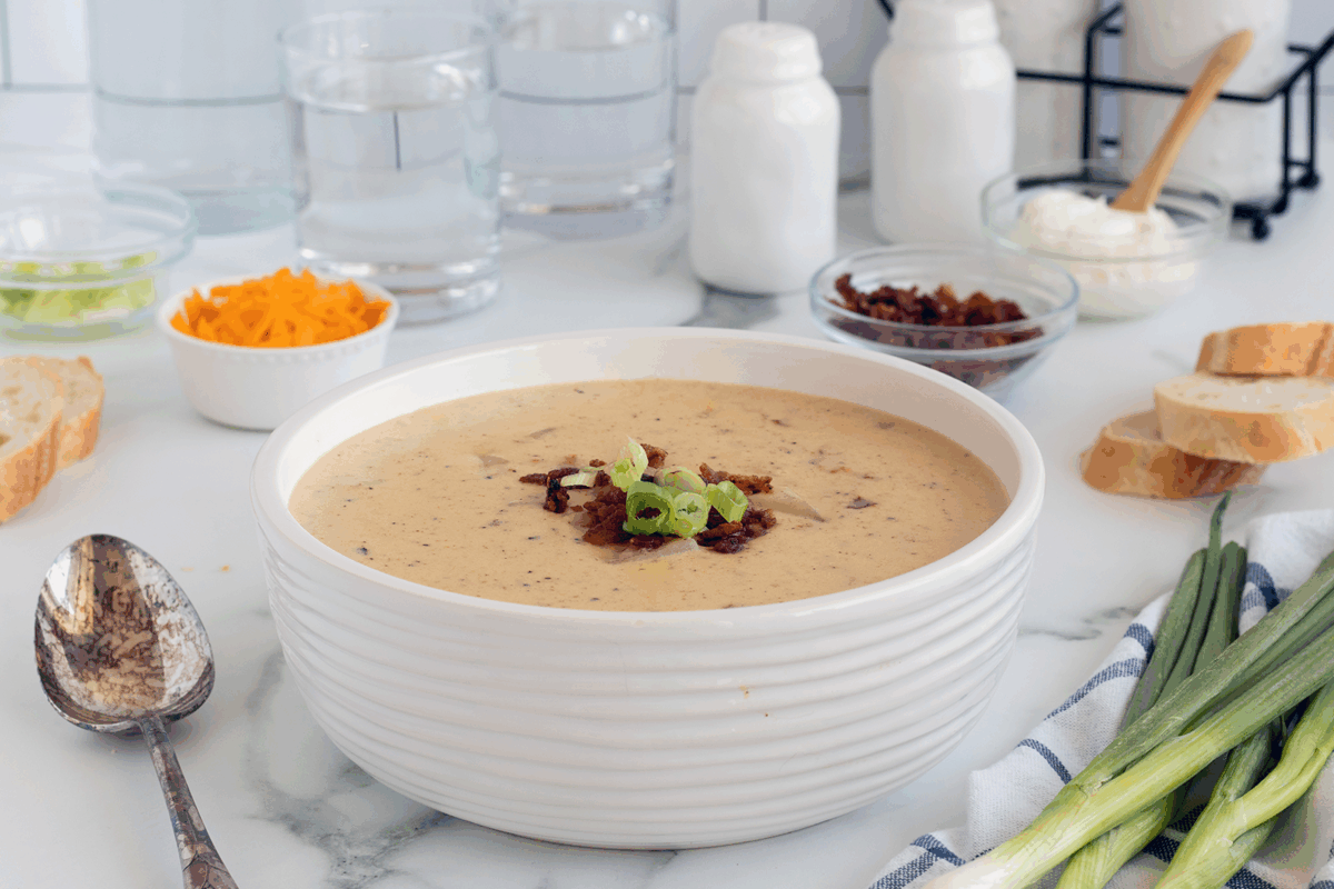 A bowl of creamy soup topped with bacon and green onions sits on a marble counter, surrounded by bread slices, cheese, sour cream, scallions, and glasses of water. A spoon and a blue-striped cloth are nearby.