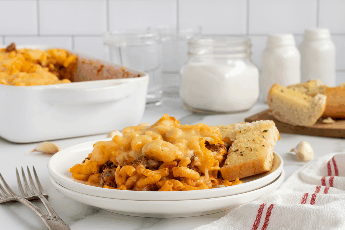 A plate of cheesy baked pasta with ground meat, served with a slice of garlic bread. In the background, there’s a baking dish of pasta, jars, glasses, and more bread on a cutting board. Forks and a towel are beside the plate.