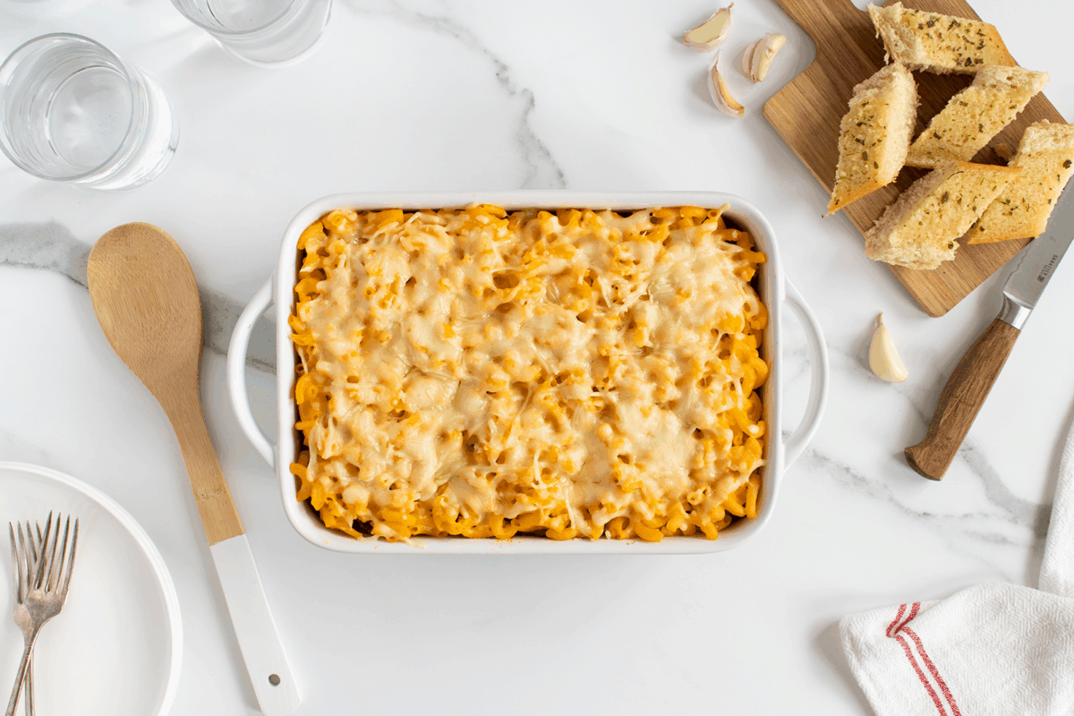 A baking dish of cheesy baked macaroni and cheese sits on a marble counter, surrounded by garlic bread slices, two glasses of water, a wooden spoon, a fork and knife, and a red-striped kitchen towel.