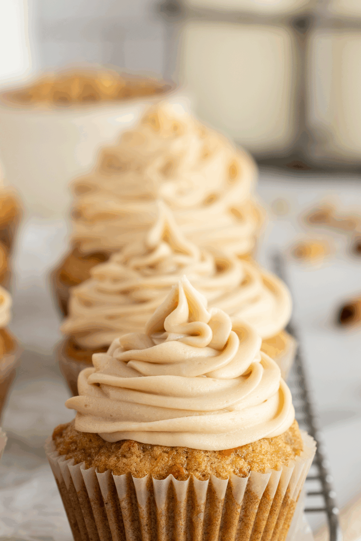 A close-up of several cupcakes with light brown swirled frosting, arranged in a row on a cooling rack, with a blurred background.