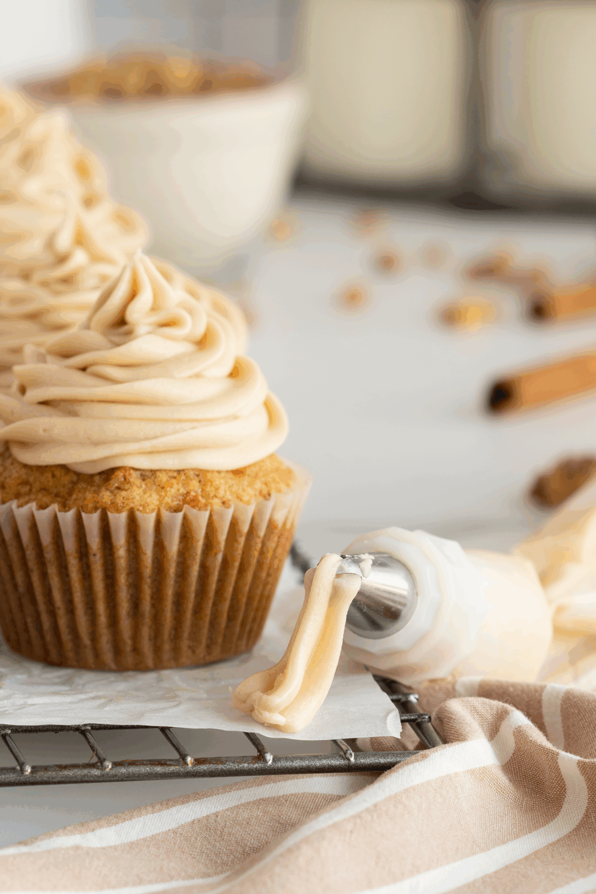 A close-up of a cupcake with swirled frosting on a cooling rack next to a piping bag with more frosting. Other cupcakes, a bowl, and cinnamon sticks are blurred in the background.