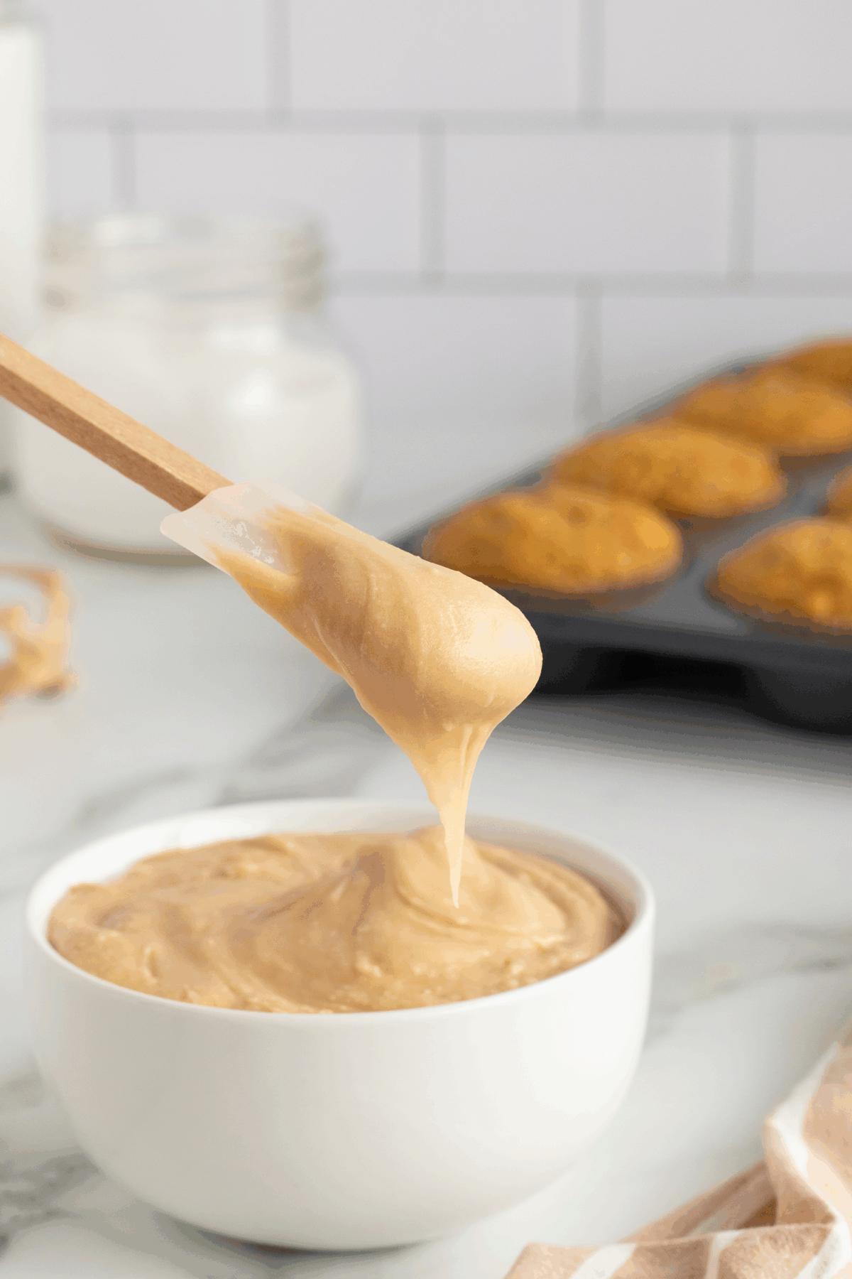 A spoon holds up thick, creamy frosting above a white bowl filled with the same frosting. In the background, a muffin tray with baked goods and a jar are visible on a marble countertop.