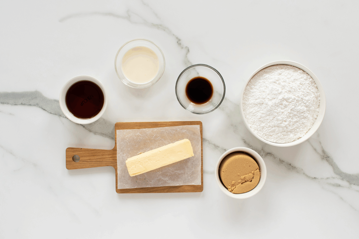 Six baking ingredients on a white marble surface: a stick of butter on a wooden board, bowls of brown sugar and powdered sugar, a small bowl of heavy cream, and two bowls with vanilla and dark liquid.