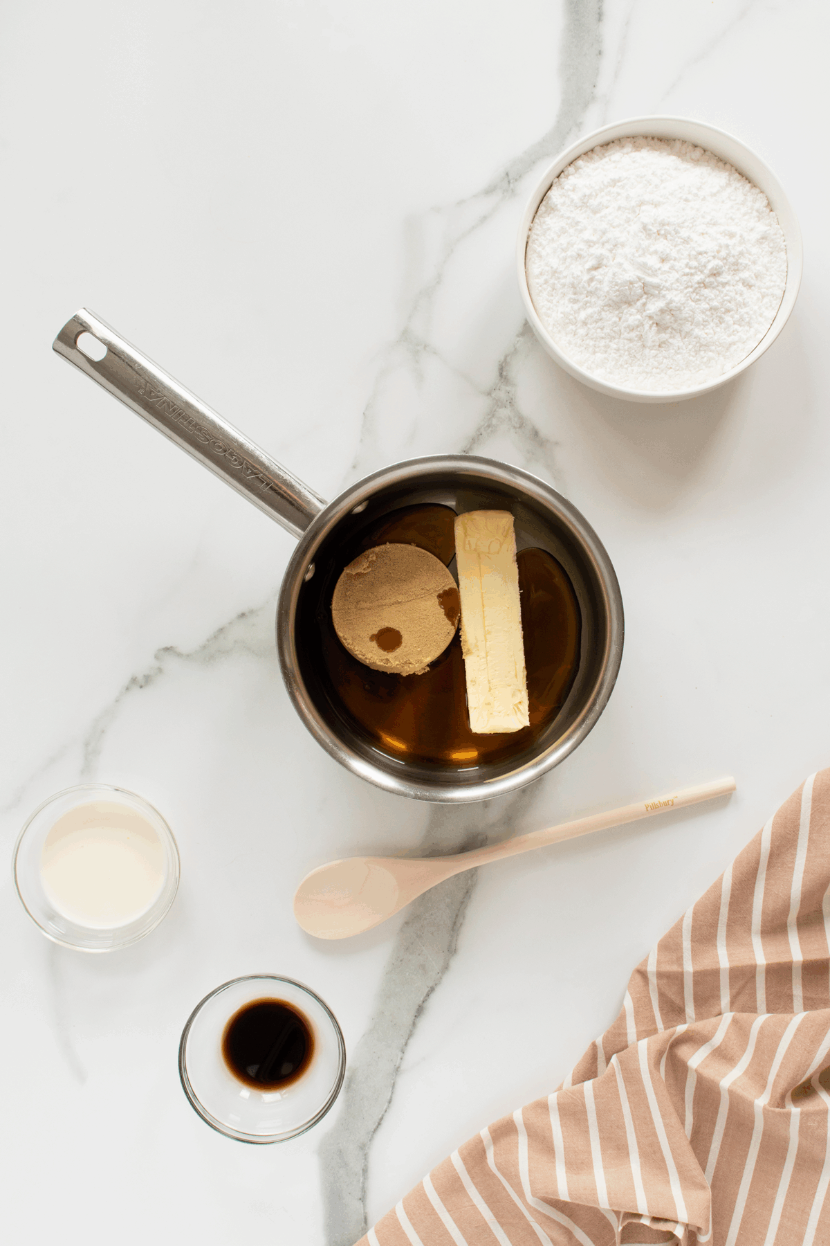 A saucepan with brown sugar, butter, and syrup, surrounded by a bowl of powdered sugar, a small bowl of cream, a bowl of vanilla, a wooden spoon, and a striped cloth on a white marble surface.