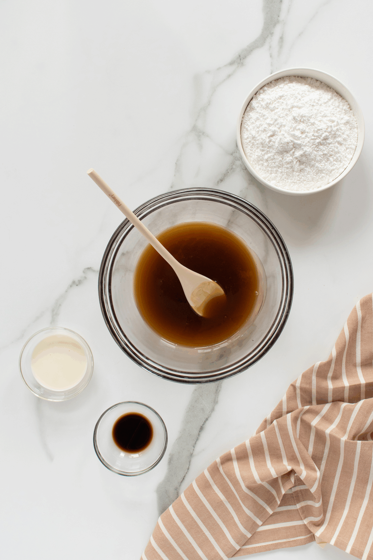 A bowl of brown liquid with a wooden spoon, a bowl of white powder, a small glass of cream, a small dish of dark liquid, and a beige-striped cloth on a white marble surface.
