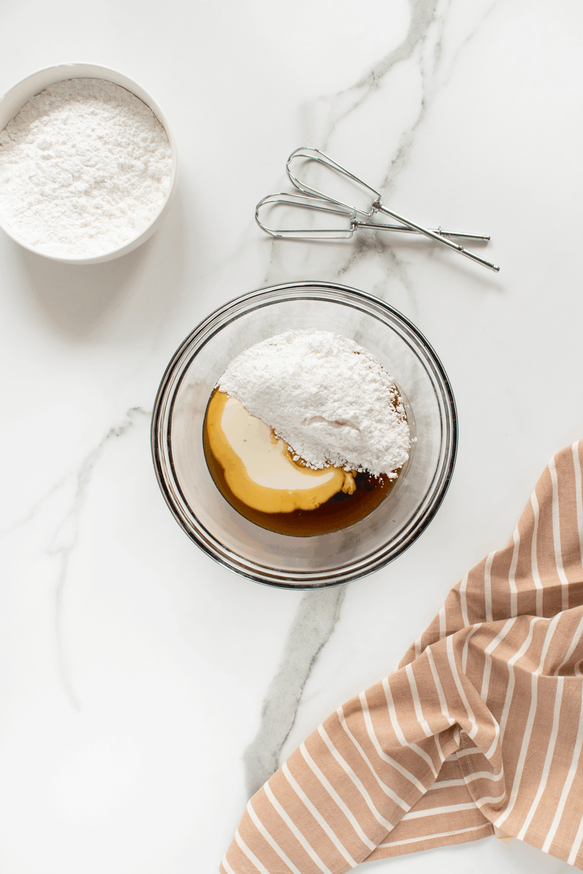 A glass bowl with powdered sugar, vanilla extract, and cream sits on a marble surface beside a bowl of powdered sugar, metal beaters, and a beige-striped cloth.