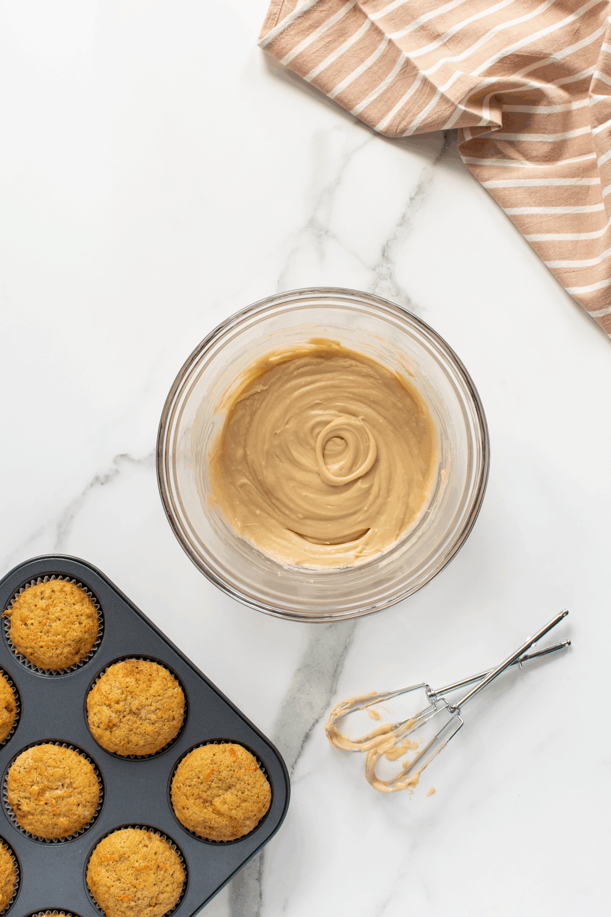 A bowl of creamy frosting sits on a marble countertop next to a pan of baked cupcakes. Two metal beaters with frosting rest nearby, and a tan striped towel is partially visible in the corner.