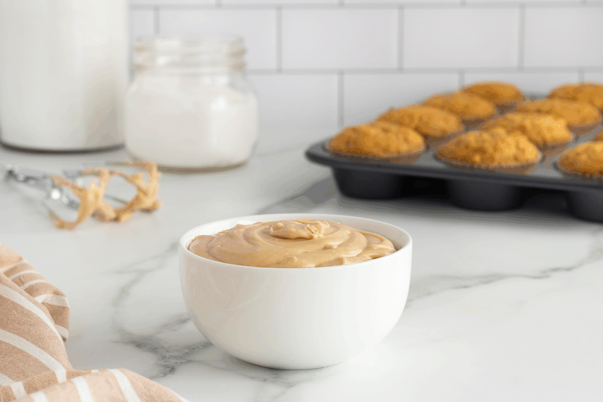 A white bowl filled with creamy peanut butter sits on a marble countertop, with a striped towel nearby, a jar in the background, and a muffin tin full of baked muffins.