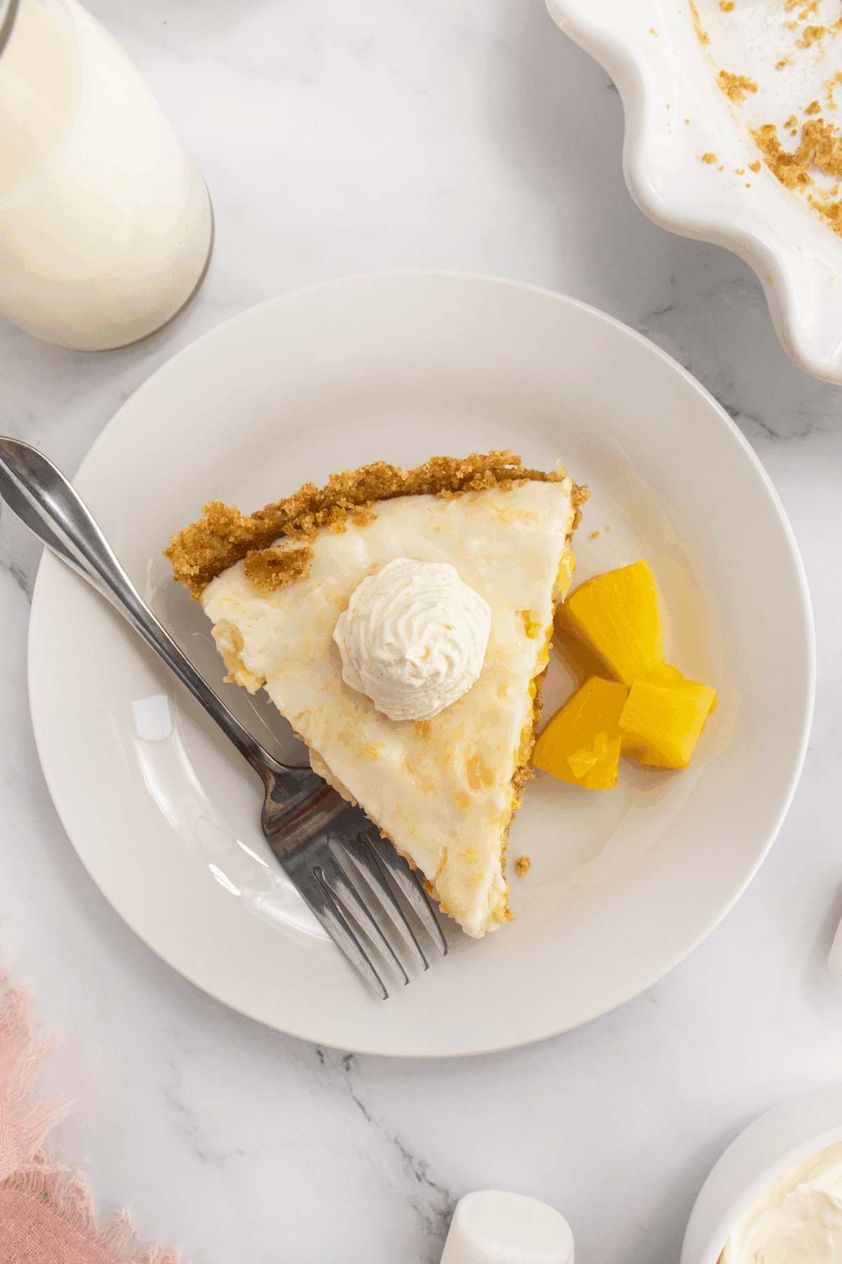 A slice of creamy pie with a graham cracker crust and a dollop of whipped cream, served with mango chunks on a white plate with a fork. A glass of milk and pie dish are nearby on a marble surface.