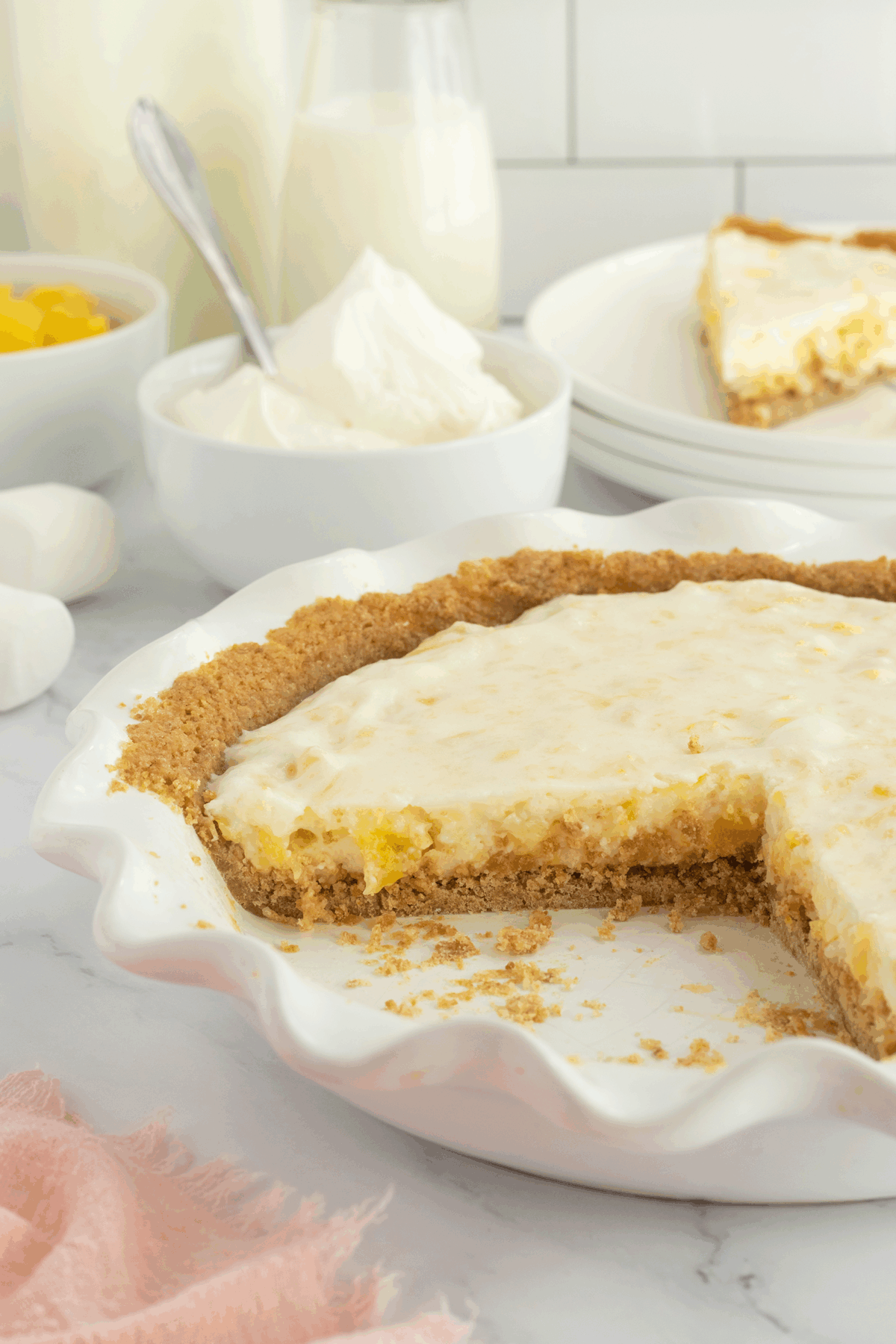 A creamy pie with a graham cracker crust sits in a white pie dish, with a slice missing. Bowls of whipped topping and fruit, a glass of milk, and stacked plates are in the background on a marble surface.
