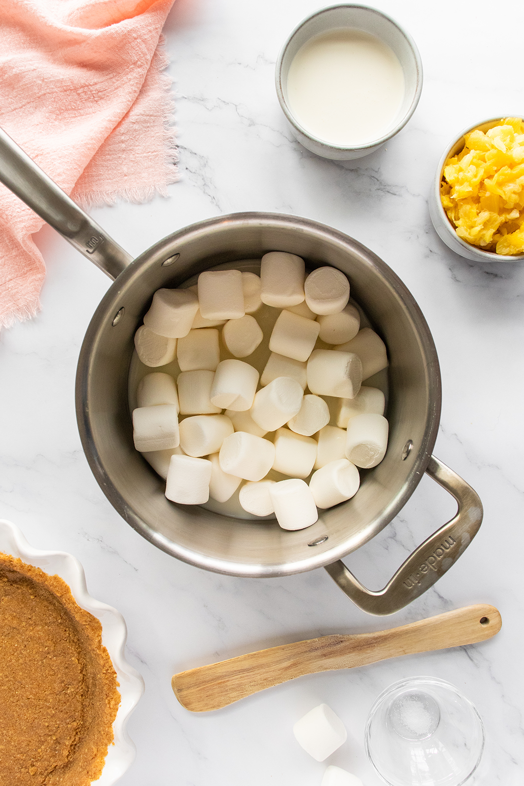 A saucepan filled with marshmallows sits on a marble surface, surrounded by a graham cracker crust in a pie dish, a wooden spatula, a bowl of crushed pineapple, a small cup of milk, and a pink cloth.