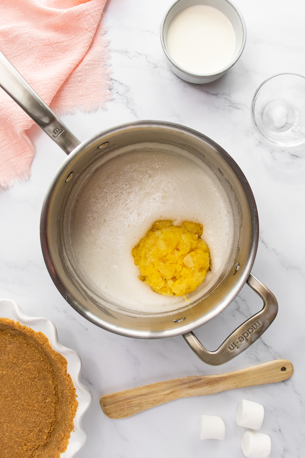 A saucepan contains a white, foamy mixture with a dollop of crushed pineapple in the center. Nearby are a wooden spatula, a pie crust, a pink towel, marshmallows, a glass, and a cup of milk on a marble surface.