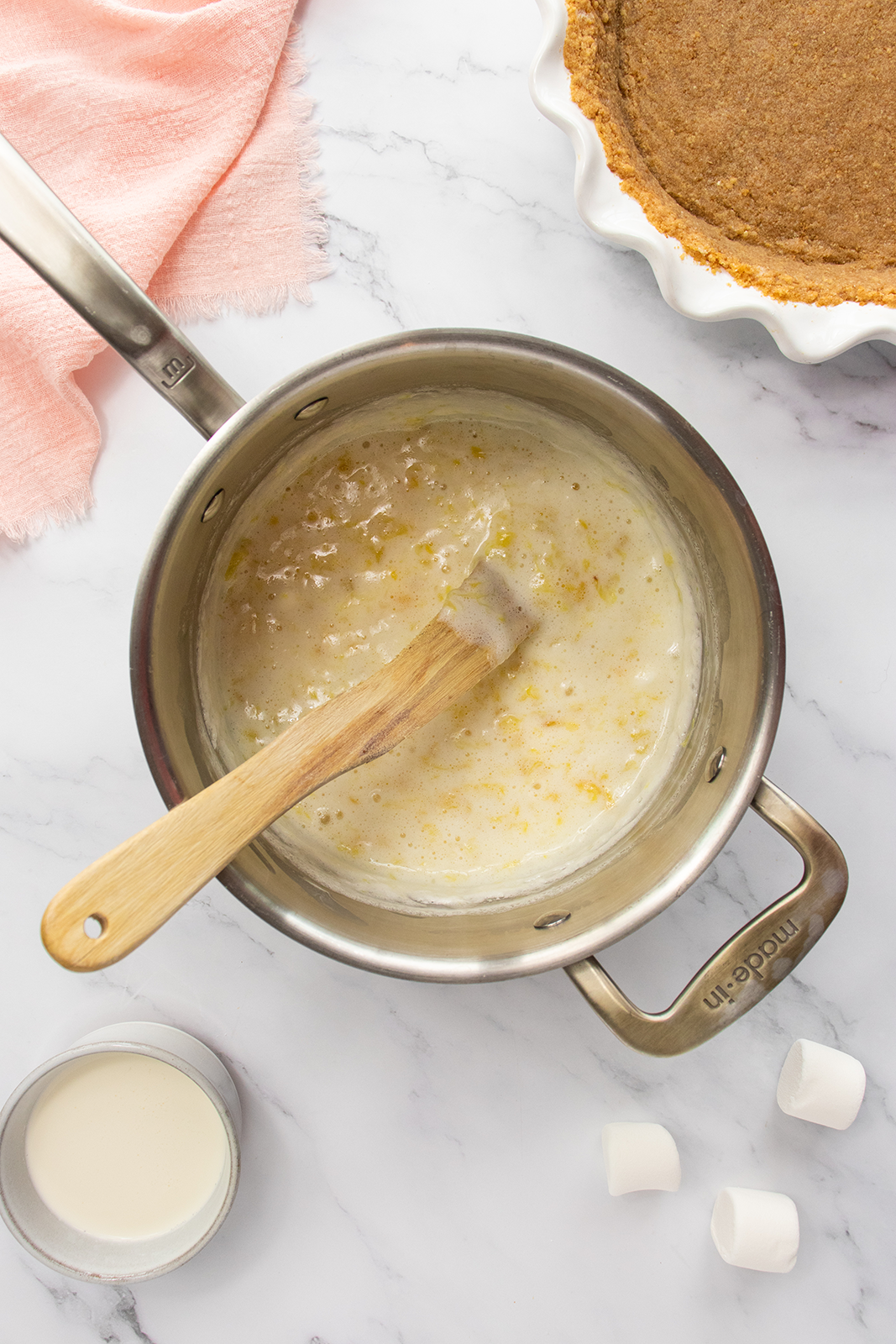 A saucepan with a wooden spoon stirring a creamy mixture sits on a marble surface, next to a cup of milk, marshmallows, a pie crust, and a pink cloth.