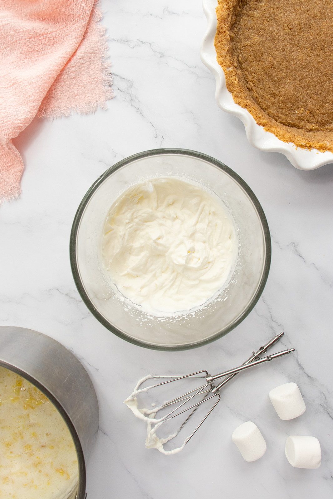 A glass bowl of whipped cream sits on a marble countertop next to a pie, a saucepan with mixture, two electric mixer beaters, marshmallows, and a pink cloth.