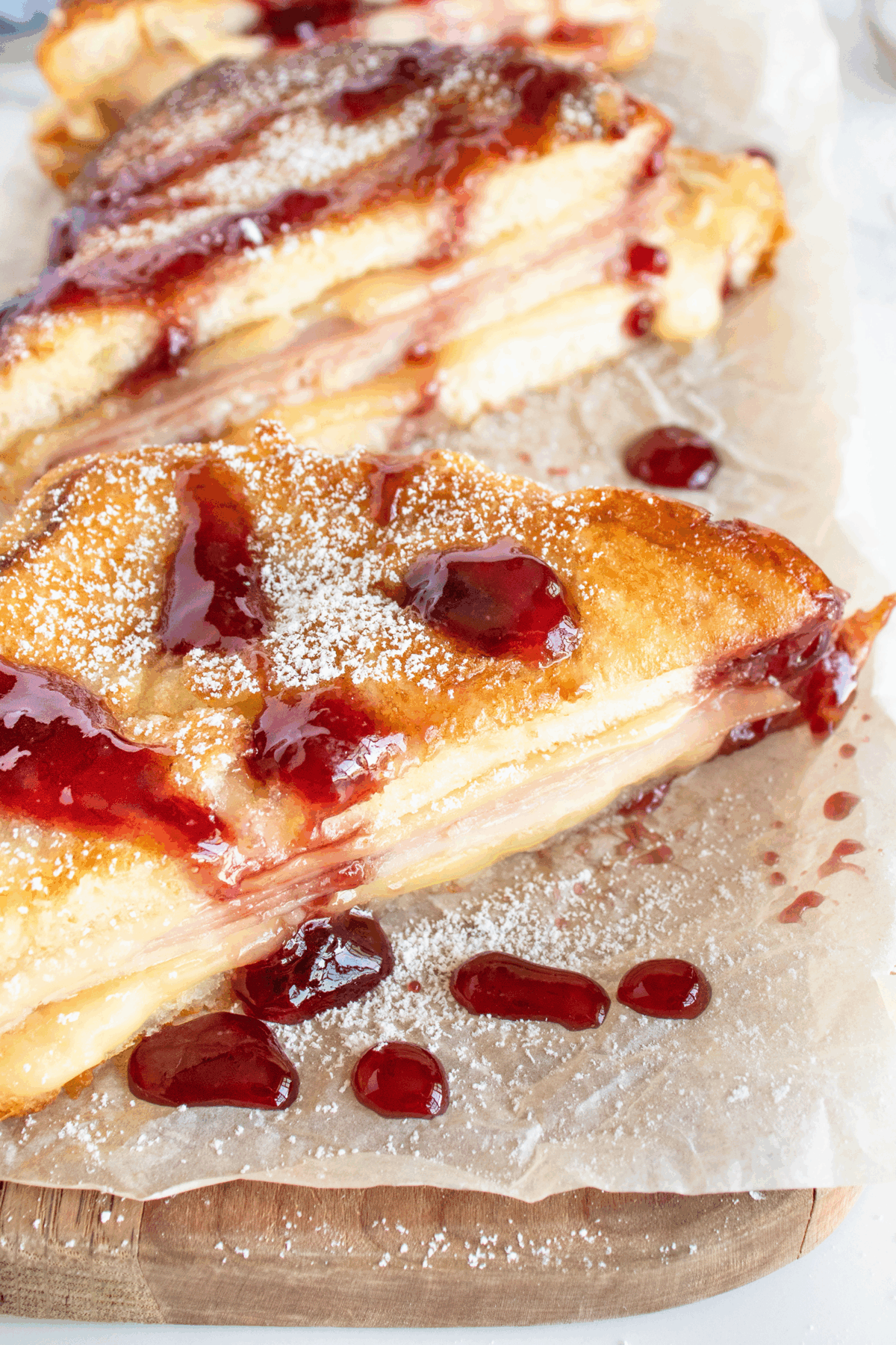 A close-up of stacked slices of Monte Cristo sandwich with layers of ham, cheese, and bread, topped with powdered sugar and drizzled with red fruit jam, served on parchment paper.