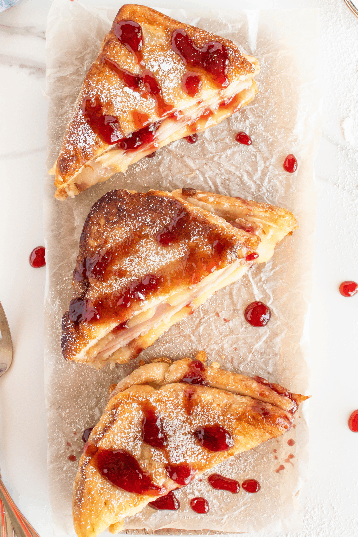 Three triangular slices of pastry dusted with powdered sugar, drizzled with red fruit sauce, arranged on parchment paper atop a white surface.