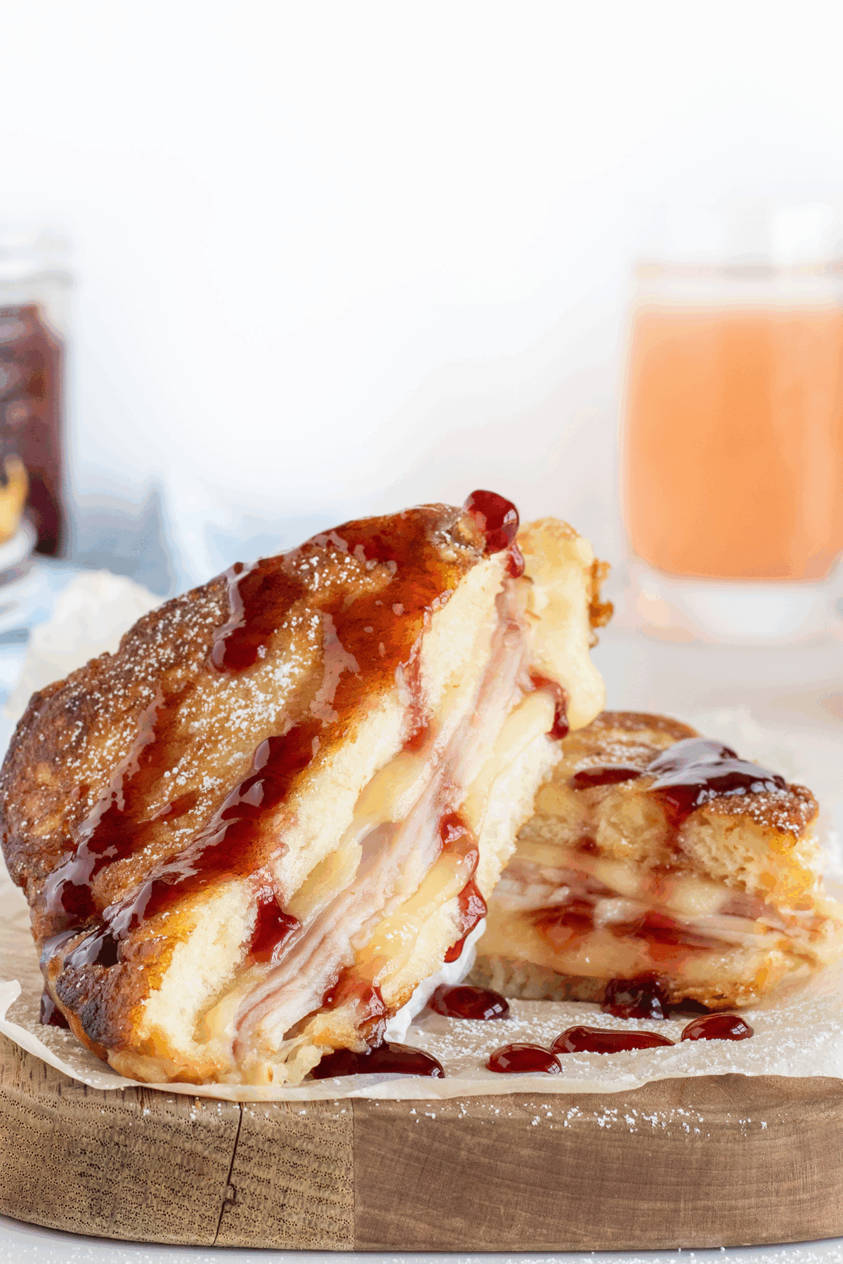 A close-up of a Monte Cristo sandwich cut in half, showing layers of ham, turkey, and cheese inside crispy bread, topped with powdered sugar and drizzled with red jam. A glass of orange drink sits in the background.