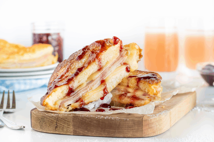 A Monte Cristo sandwich cut in half, filled with turkey, ham, and melted cheese, dusted with powdered sugar, and topped with berry jam, sits on a wooden serving board with drinks and jam jars in the background.