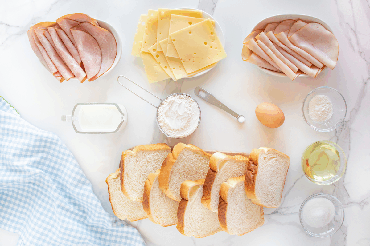 Slices of bread, ham, turkey, Swiss cheese, an egg, flour, milk, oil, powdered sugar, and a measuring spoon arranged on a white marble surface with a blue gingham cloth on the side.