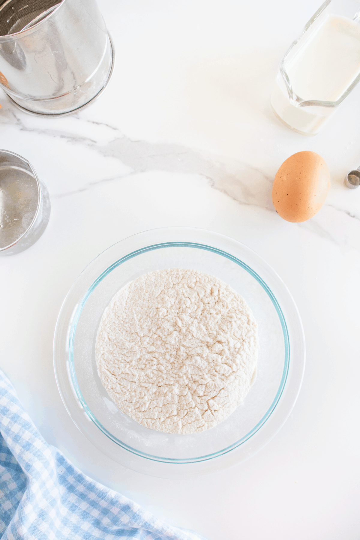 A glass bowl of flour on a white marble surface, surrounded by an egg, a measuring cup of milk, a metal sifter, and a blue checkered cloth.
