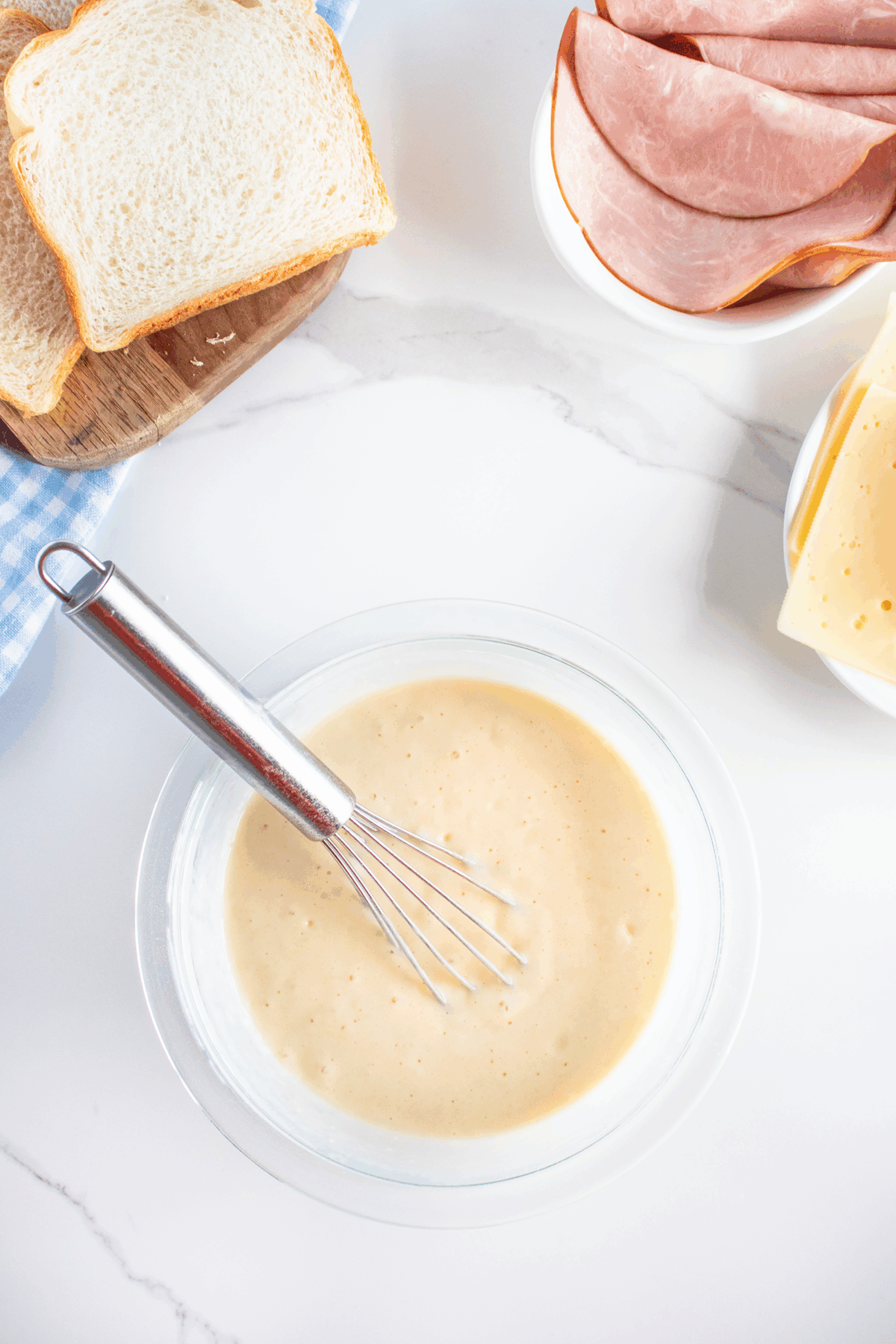 A glass bowl of creamy white sauce with a whisk inside, surrounded by sliced bread, ham, and cheese on a white marble surface.