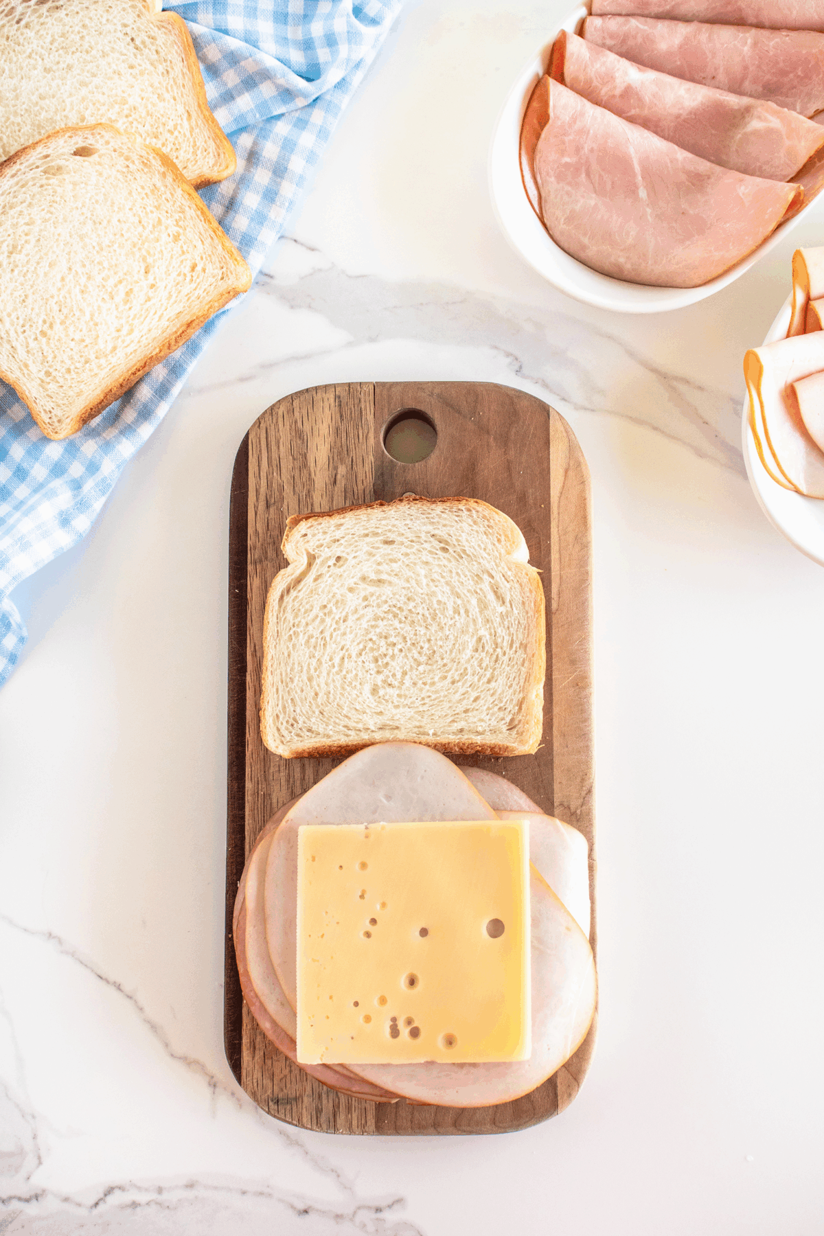 Two slices of white bread, a slice of ham, turkey, and Swiss cheese on a wooden board. Nearby are more slices of bread, ham, and turkey on a white marble counter with a blue checkered cloth.
