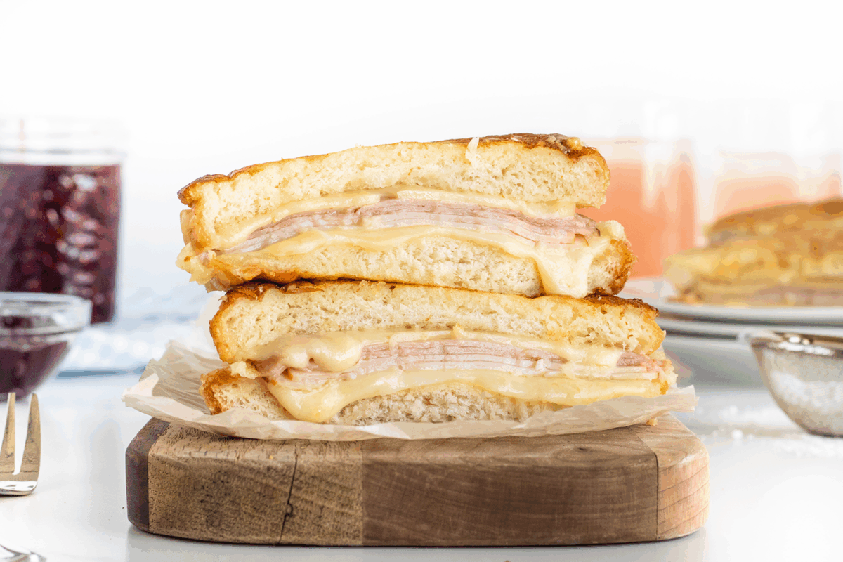 A close-up of a stacked ham and cheese sandwich, cut in half to show melted cheese and layers of ham, placed on a wooden board with blurry jars and drinks in the background.