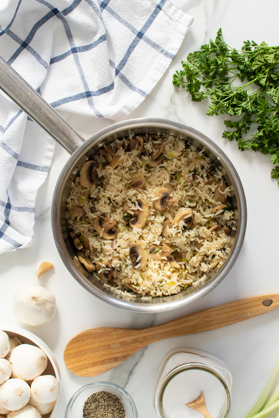 A saucepan filled with cooked rice and sliced mushrooms sits on a white marble surface, surrounded by fresh parsley, a wooden spoon, garlic, mushrooms, jars of salt and pepper, and a checkered kitchen towel.