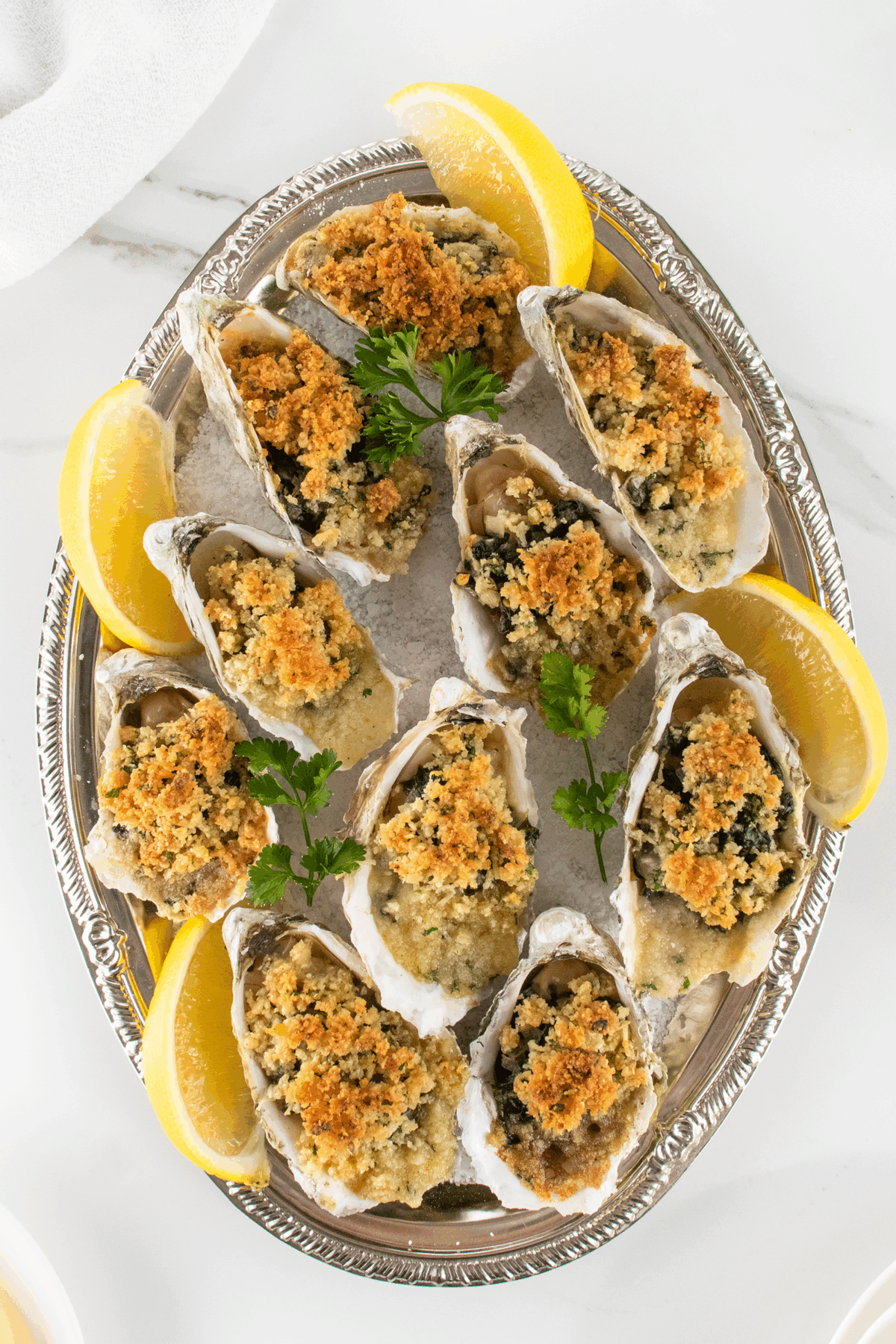 A silver platter with baked oysters topped with golden breadcrumbs, garnished with parsley and lemon wedges, set on a white marble surface.