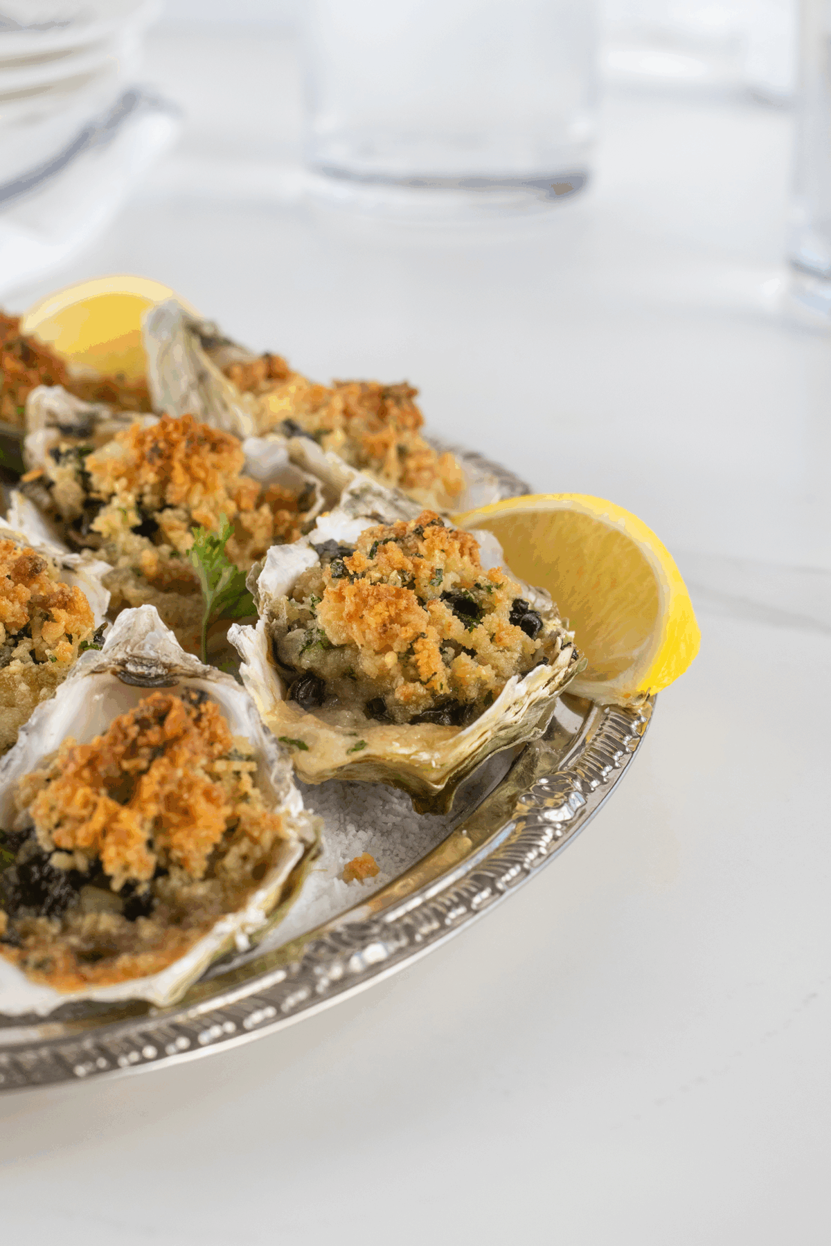 A silver platter of baked oysters topped with golden breadcrumbs, garnished with lemon wedges, sits on a white surface. The oysters appear crispy and freshly prepared.